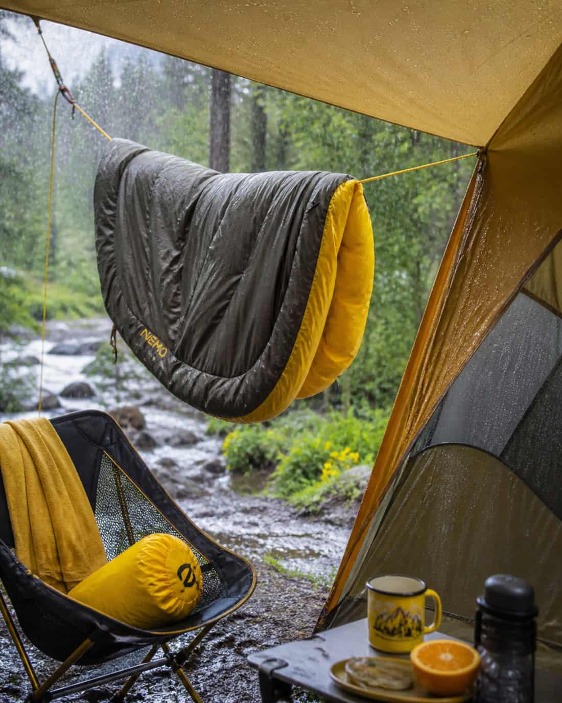 A sleeping bag hangs under a campsite tarp to air out during rain, with visible tent condensation, wet forest surroundings, and bright citrus-yellow accents creating an upbeat mood